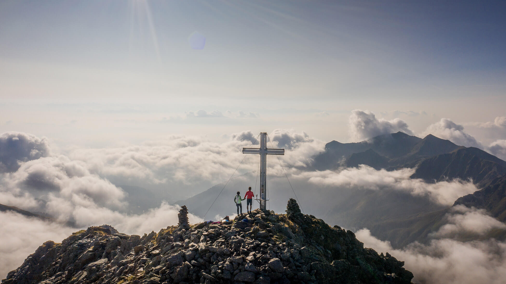 Zwei Wanderer stehen neben einem großen Kreuz auf einem Berggipfel, umgeben von Wolken und fernen Bergen. - Bucherhof