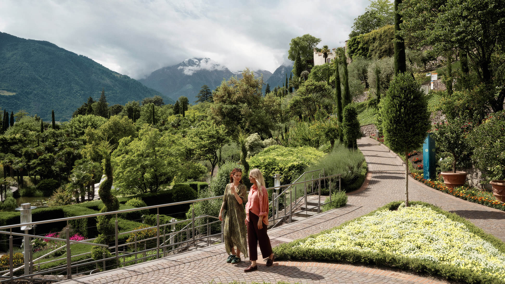 Zwei Frauen spazieren auf einem Gartenweg, umgeben von üppigem Grün und mit den Bergen im Hintergrund. - Bucherhof