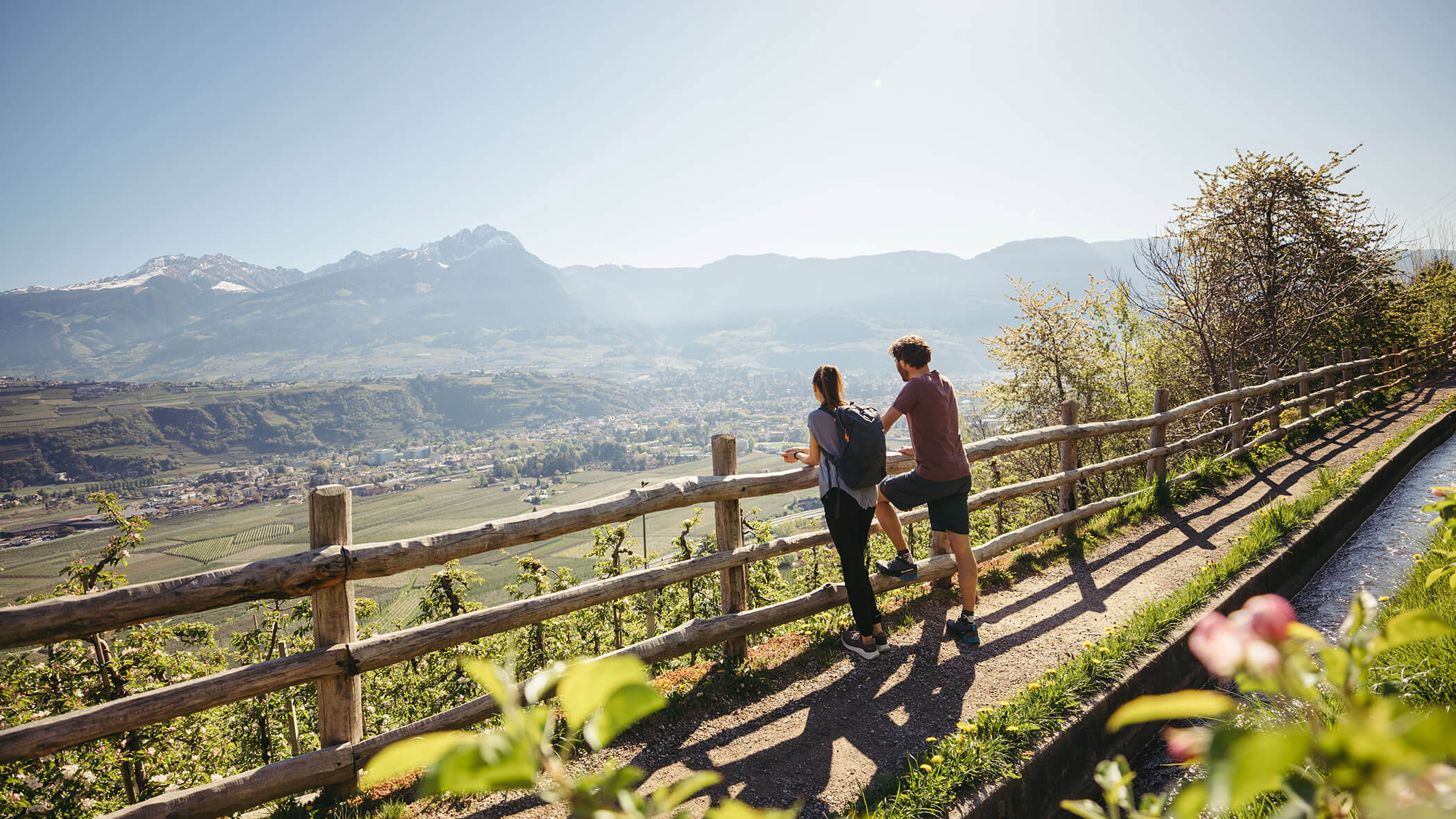 Zwei Menschen stehen an einem Holzzaun und blicken auf ein malerisches Tal mit Bergen im Hintergrund. - Bucherhof
