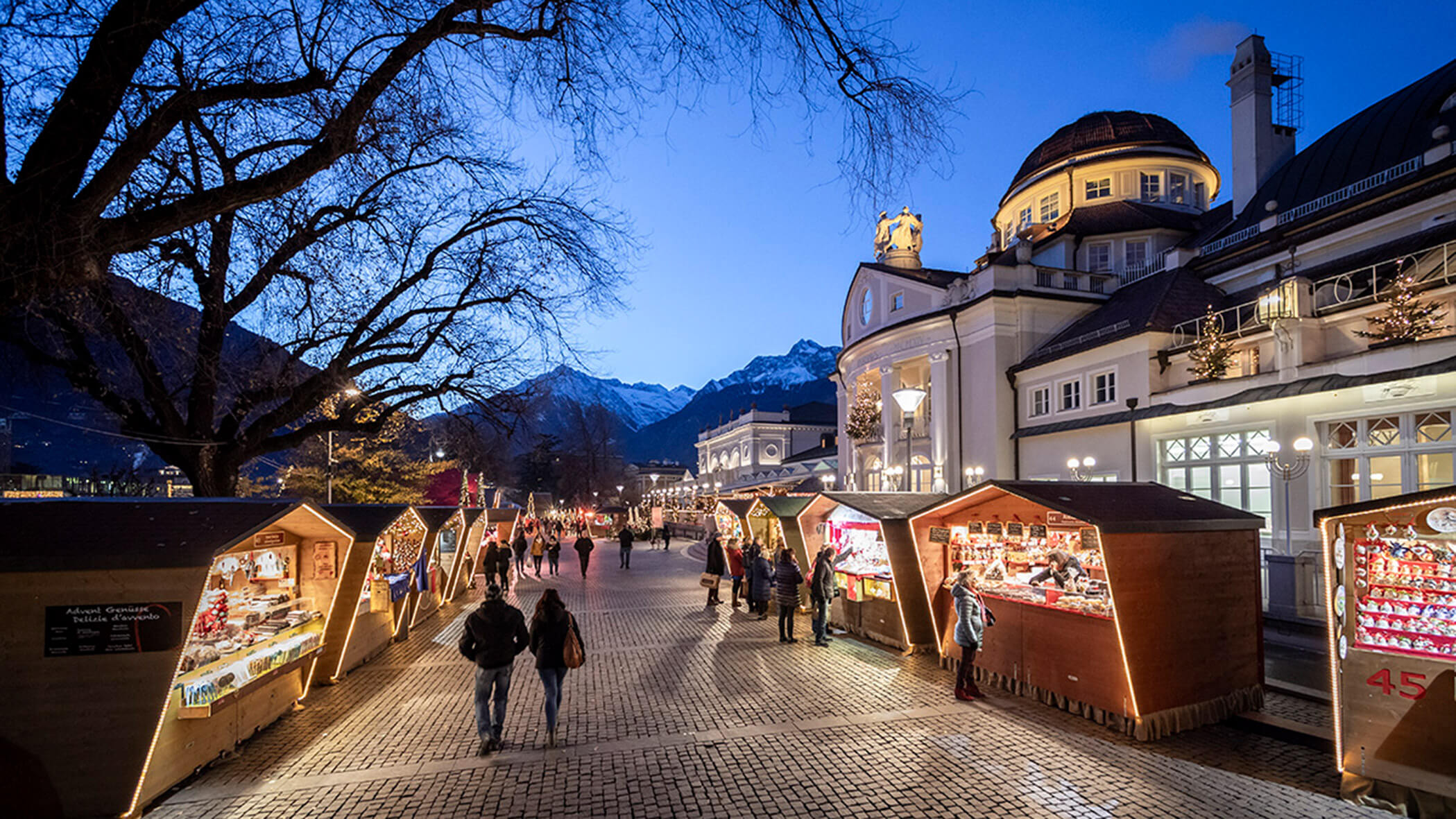 Menschen, die in der Abenddämmerung an einem Markt im Freien an Holzständen stöbern, im Hintergrund sind Berge und Gebäude zu sehen. - Bucherhof