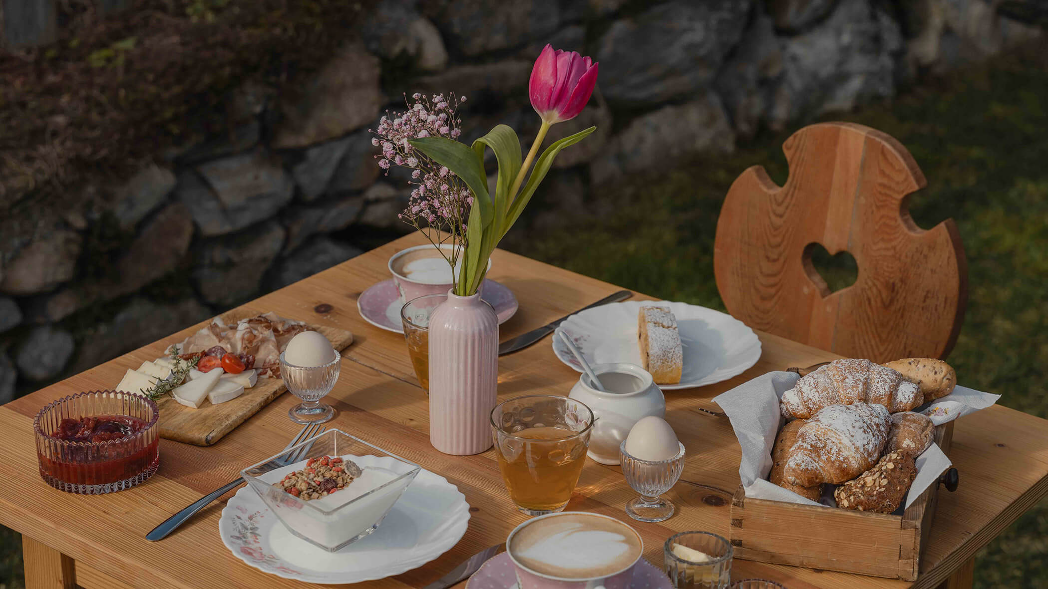 Ein Holztisch im Freien, gedeckt mit Gebäck, Eiern, Getränken und einer Vase mit rosa Blumen. - Bucherhof