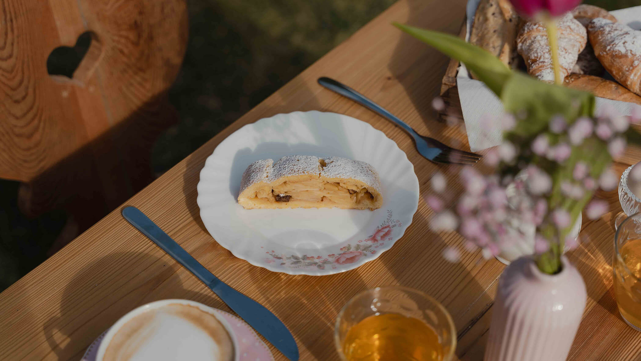 Ein Stück Apfelstrudel auf einem weißen Teller, der auf einem Holztisch steht, zusammen mit Getränken, Blumen und Besteck. - Bucherhof