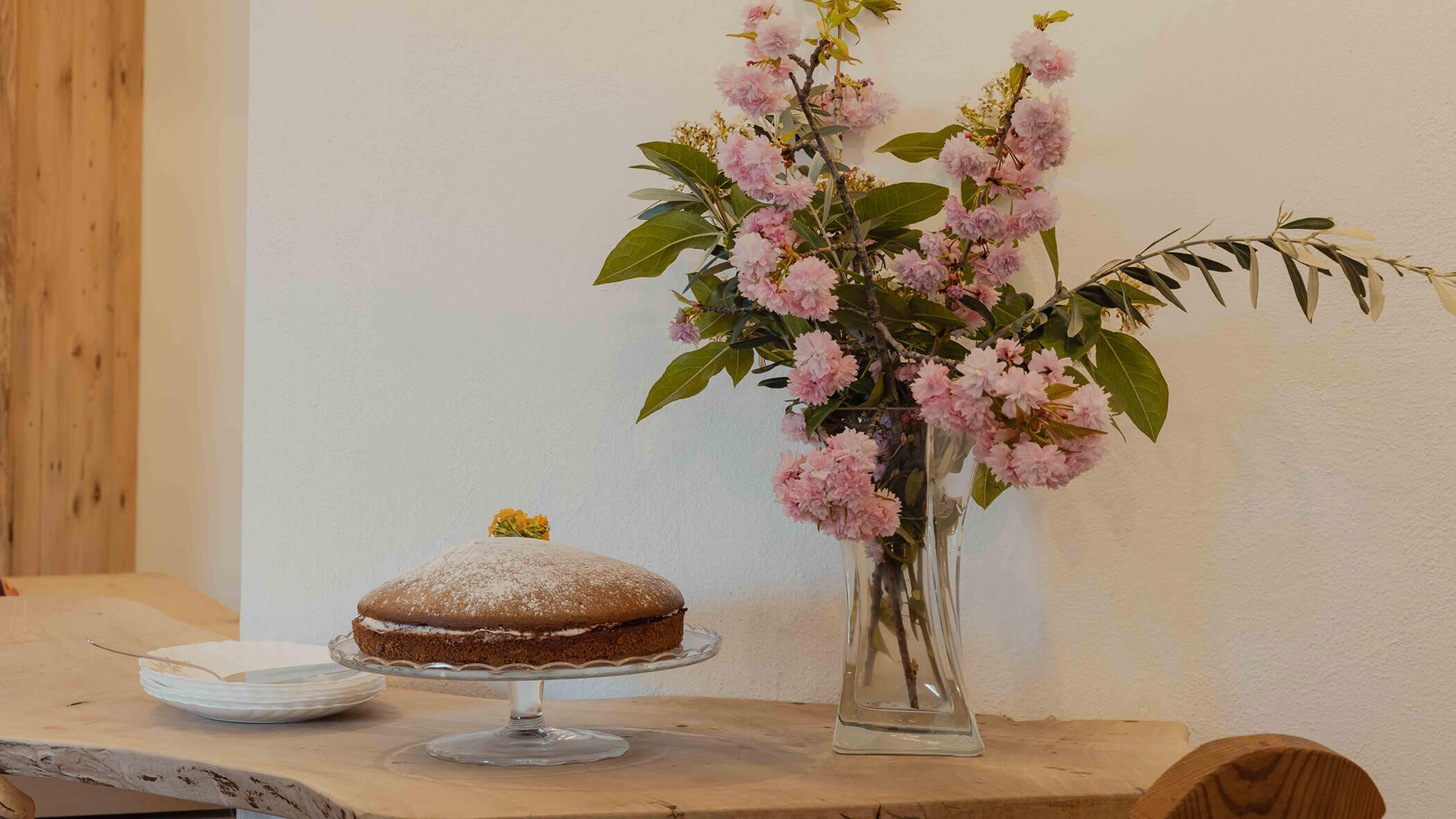 Ein runder Kuchen auf einem Glasständer neben einer Vase mit rosa Blumen auf einem Holztisch vor einer weißen Wand. - Bucherhof