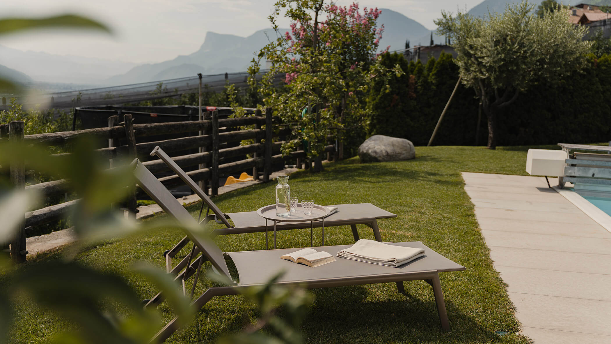 Zwei Liegestühle am Pool mit einem Buch, Handtüchern und Getränken, inmitten eines Gartens mit Blick auf die Berge. - Bucherhof