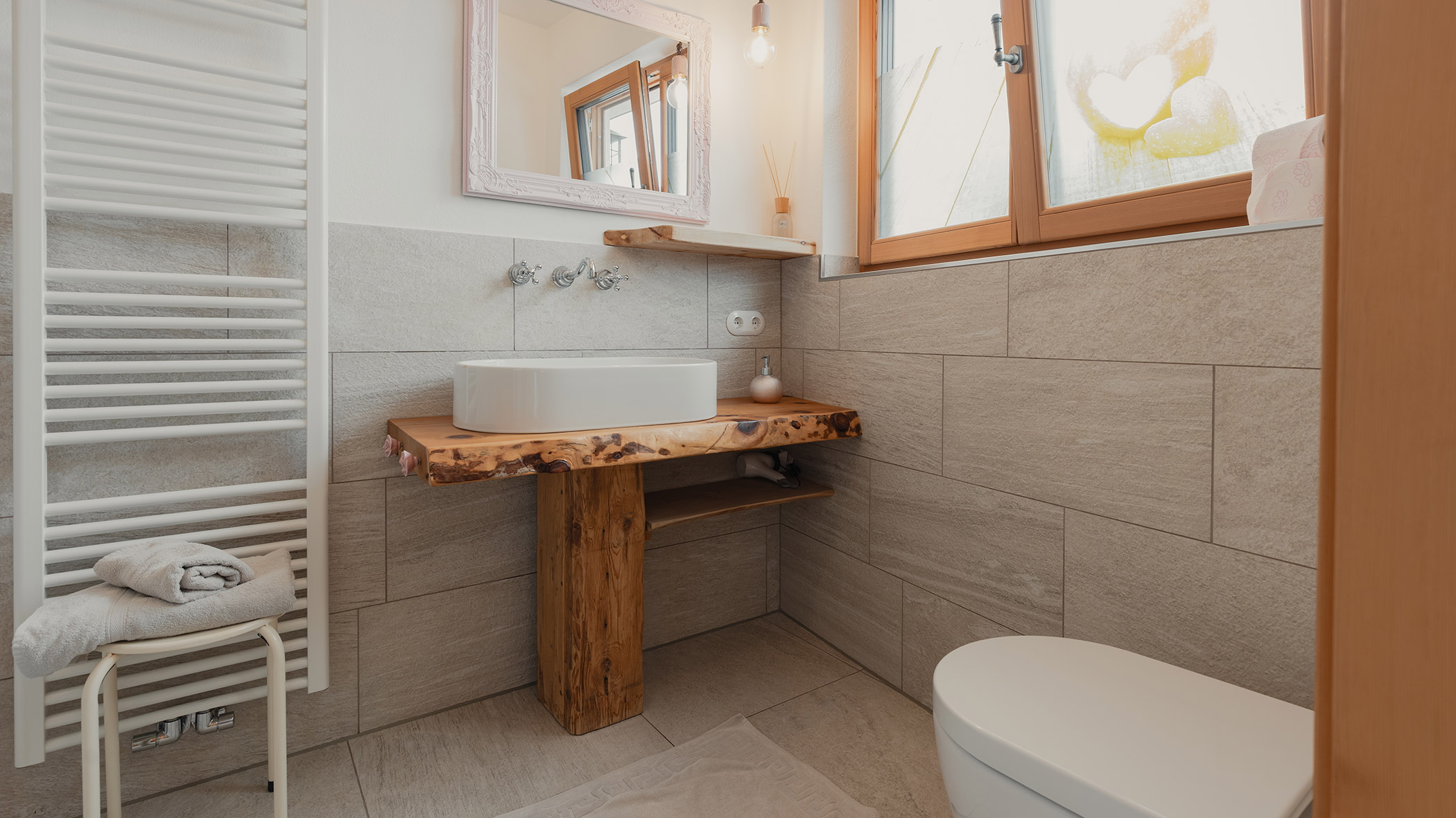 Modernes Badezimmer mit einem Waschtisch aus Holz, einem runden Waschbecken, einem Handtuchhalter, einem Fenster und großen beigen Fliesen. - Bucherhof
