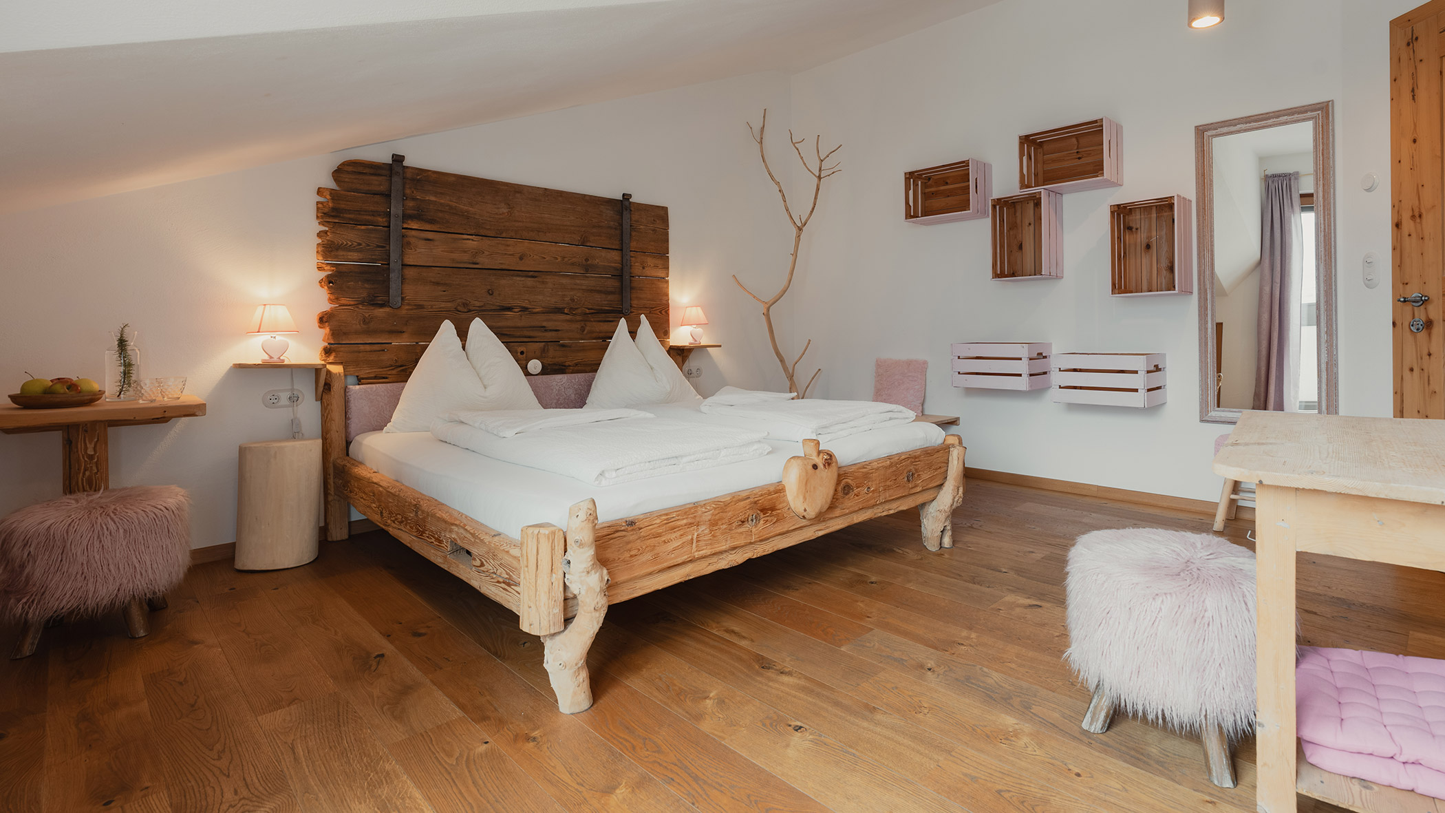 Gemütliches Schlafzimmer mit einem rustikalen Holzbett, Regalen aus Kisten und zartrosa, plüschigen Hockern auf einem Holzboden. - Bucherhof