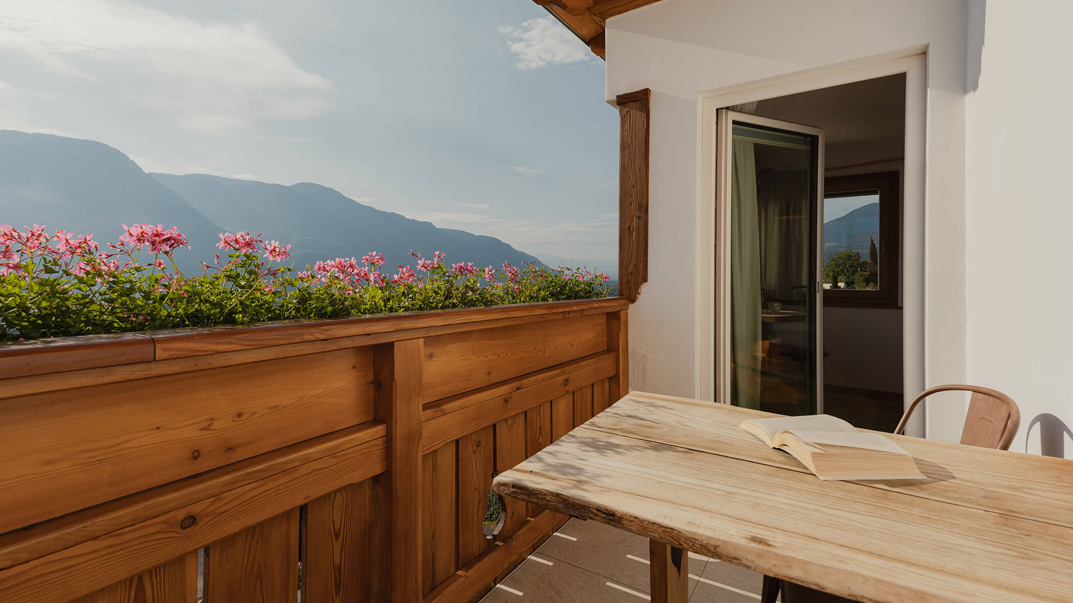 Ein Holzbalkon mit Blumen, einem Tisch, einem aufgeschlagenen Buch und Bergen im Hintergrund unter blauem Himmel. - Bucherhof