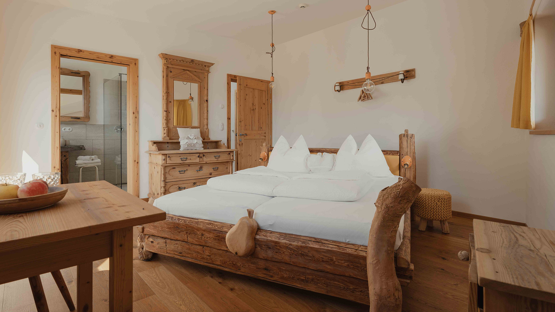 Rustikales Schlafzimmer aus Holz mit einem großen Bett, einer Kommode, einer Tür zum Badezimmer und Tageslicht, das durch das Fenster hereinströmt. - Bucherhof