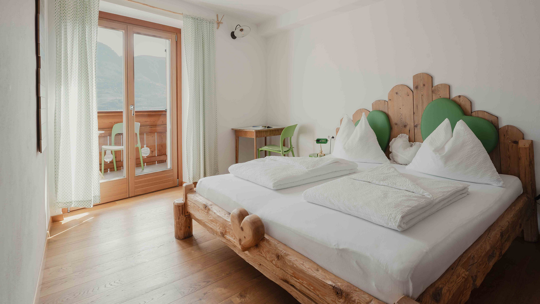 Ein sonnendurchflutetes Hotelzimmer mit einem rustikalen Holzbett, Balkontüren, einem Schreibtisch und einer Einrichtung mit grünen Akzenten. - Bucherhof