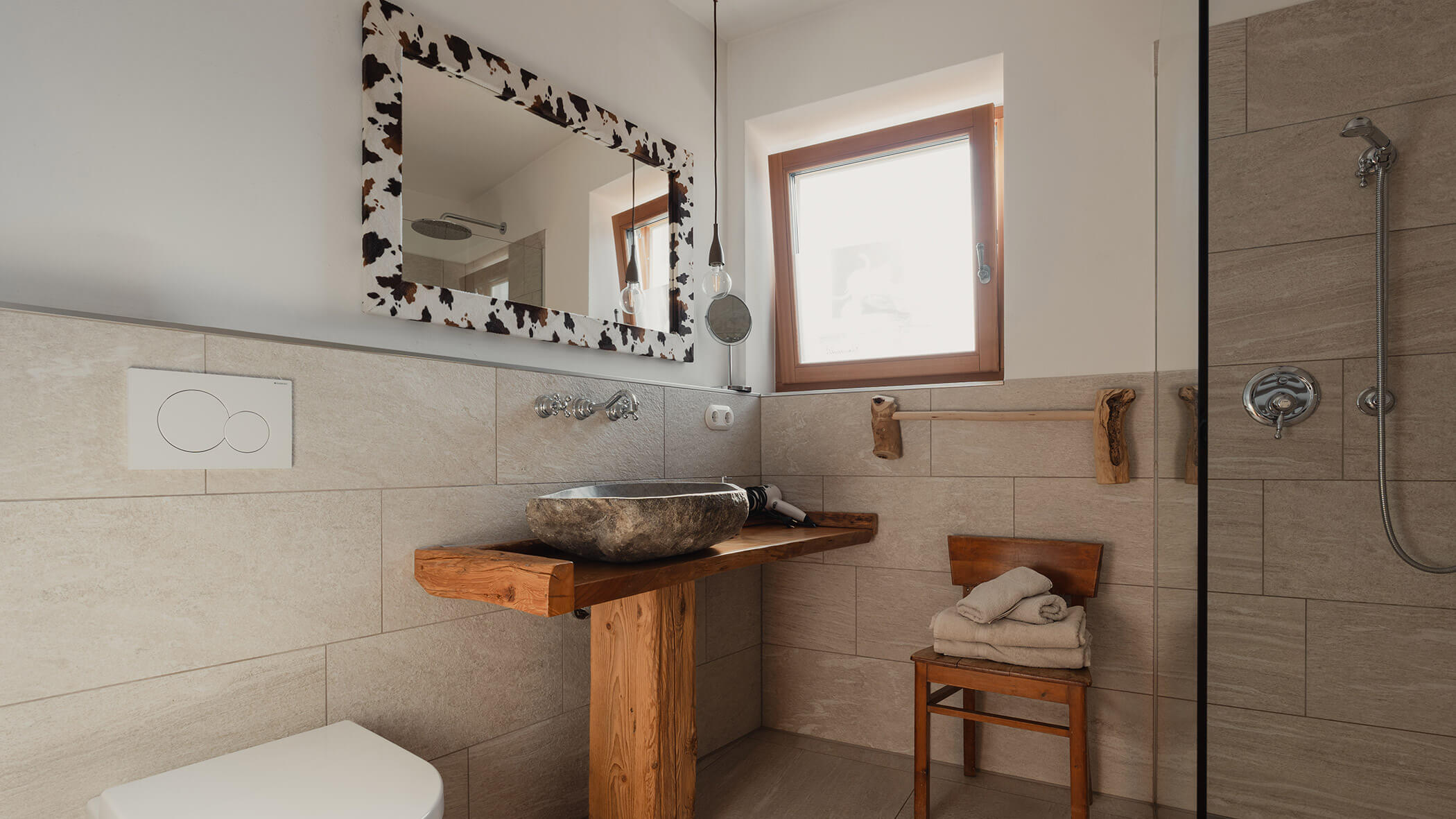 Modernes Badezimmer mit einem Waschbecken aus Stein, Holzakzenten, einem Wandspiegel, einem kleinen Fenster, einer Dusche und einem Handtuch auf einem Stuhl. - Bucherhof
