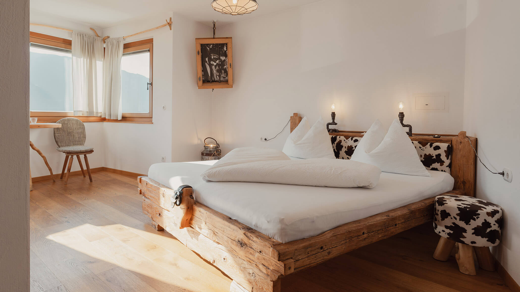 Sonniges Schlafzimmer mit einem rustikalen Holzbett, weißer Bettwäsche, einem Hocker aus Rindsleder und einem Fenster mit transparenten Vorhängen. - Bucherhof