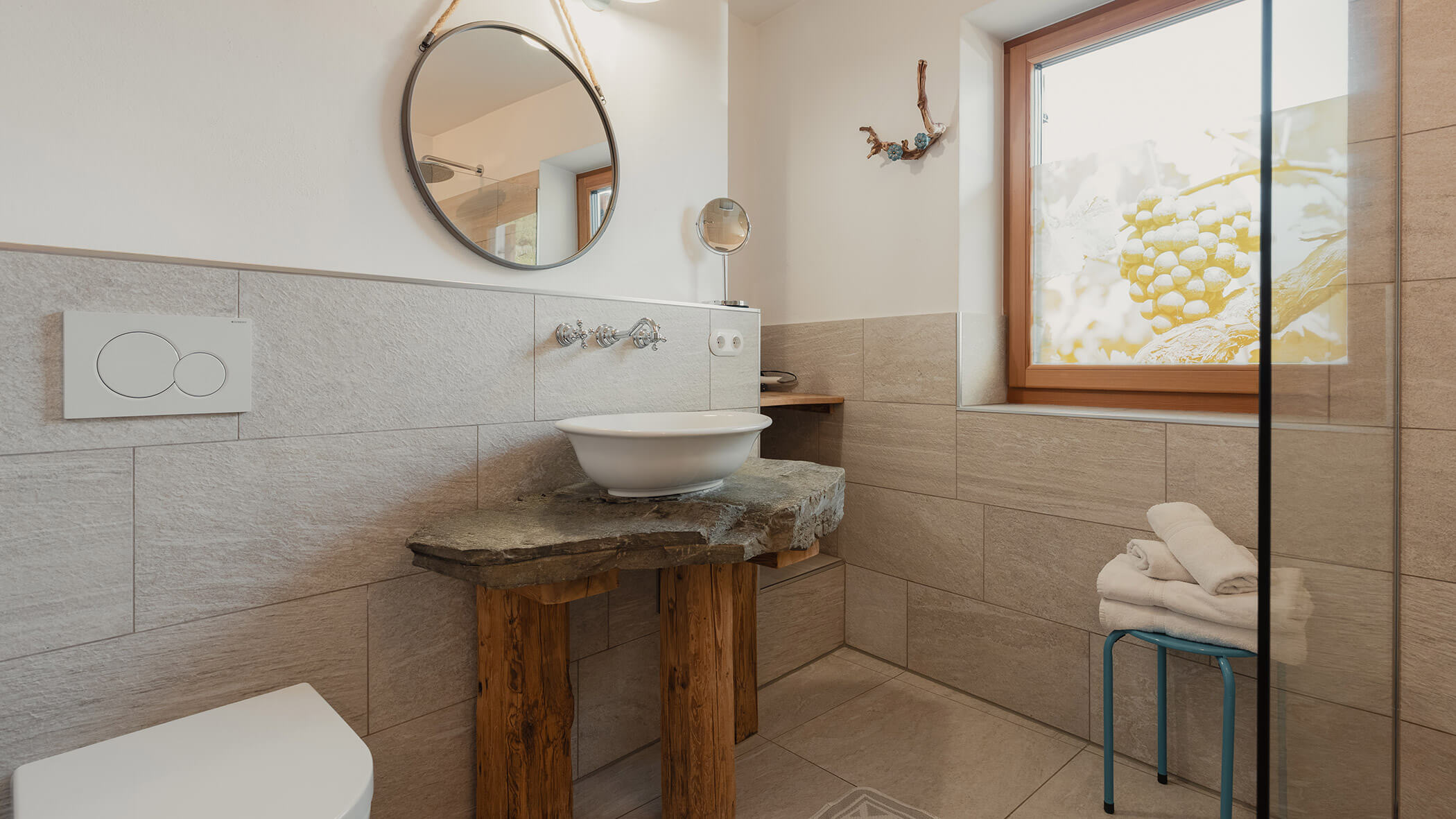 Modernes Badezimmer mit einem Waschbecken aus Stein, einem runden Spiegel, Handtüchern auf einem Hocker und einem Fenster mit einem Rollo mit Naturmotiv. - Bucherhof