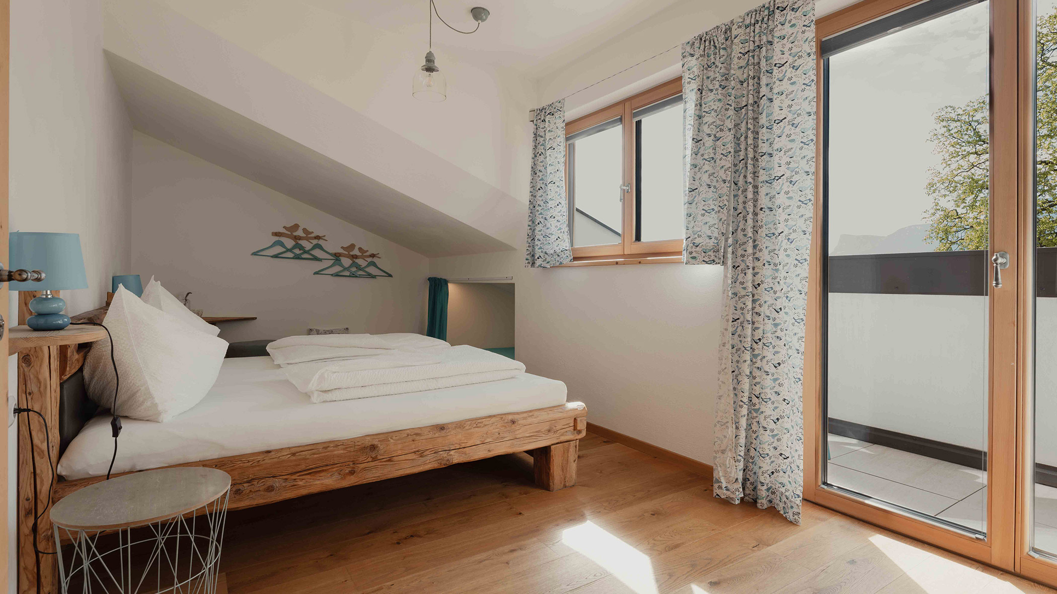 Helles Schlafzimmer mit einem Holzbett, blauen Akzenten, einem großen Fenster und einer Balkontür, durch die Tageslicht hereinströmt. - Bucherhof